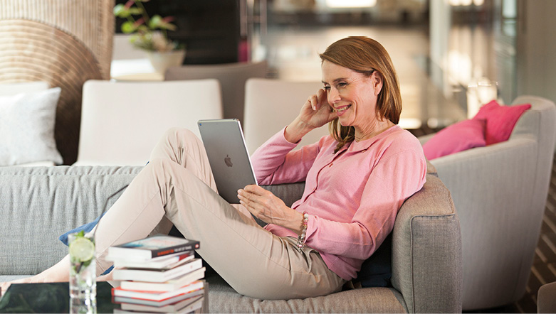 Woman holding an iPad and smiling as she reads an email while relaxing on a comfortable grey sofa