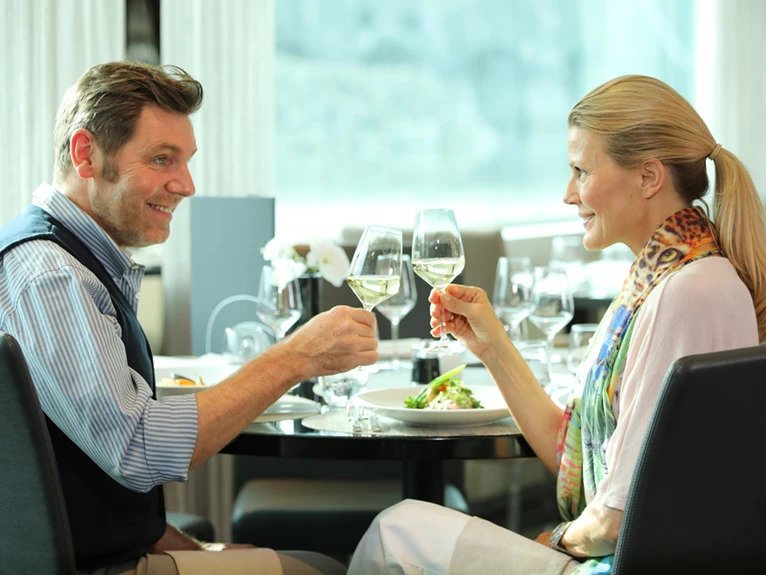 A couple toasting with glasses of wine as they enjoy a meal at the Crystal Dining restaurant on board a Scenic cruise ship.  