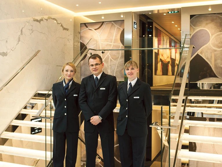 Three Scenic staff members, dressed in black suits, standing inside the elegant interior of the cruise ship.