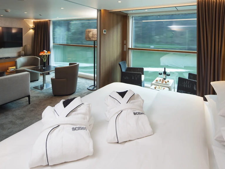 A Queen size bed adorned with plush white bathrobes in the Royal Balcony Suite, with floor-to-ceiling windows offering a view. 