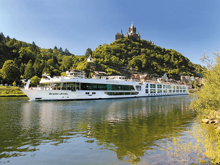 The Scenic Jewel Space-Ship gliding on a tranquil river, with a castle perched atop a hill in the background,
