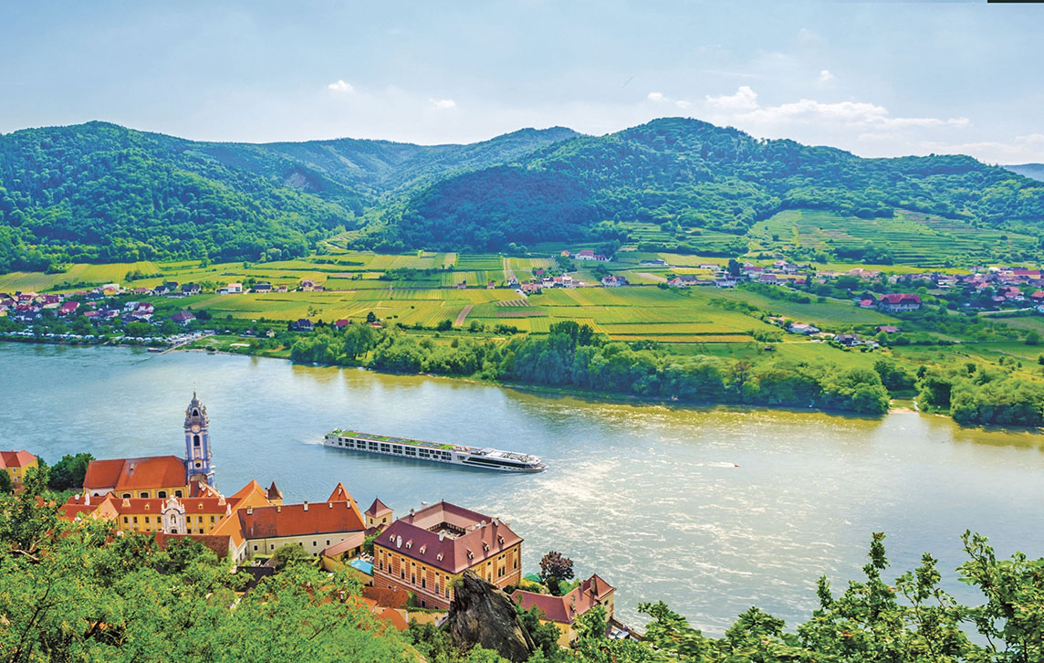 The Scenic Jasper ship cruising along the Danube River, framed by lush green fields, towering mountains, and historic buildings in Durnstein, Austria.