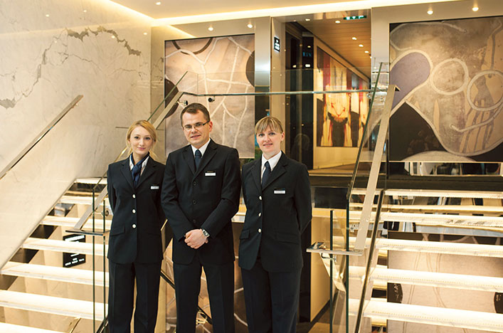Three professional crew members dressed in black pant suits, standing inside a Scenic cruise ship