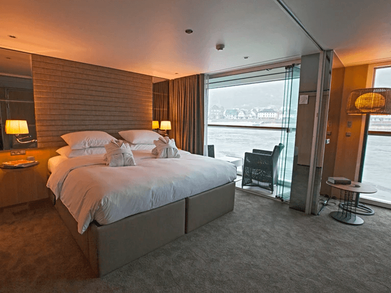 A luxurious queen bed next to floor-to-ceiling windows, with views of the passing river in the Royal Balcony Suite.