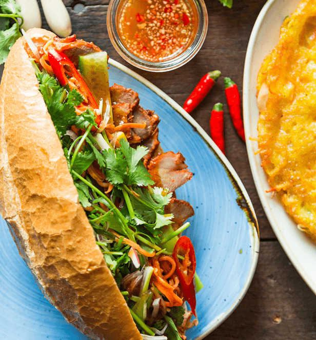 Vietnamese bánh mì sandwich with grilled pork, pickled vegetables, cilantro, and red chili on a blue plate.